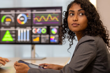Hispanic latin business woman interacting with virtual data interface. African american female observes financial data displayed on a large screen monitor, contemplating market trends and strategy.