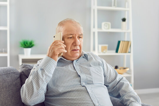 Serious senior man sits on the sofa in the home living room, talking on the phone. Engaged in a conversation, he communicates with a focused expression, highlighting the importance of the call.
