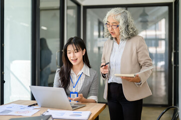 Obraz premium Old asian businesswoman holding pen and book looking at laptop with young coworker at office's table