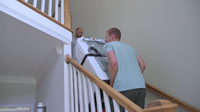 white men movers carrying washer down stairwell, coordinated lift around banister, teamwork under indoor daylight, strap and harness support, careful communication and steady footing