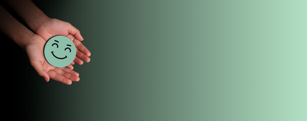 Hands gently holding a green smiling face cutout symbolizing positivity, emotional care, and mental wellness on a clean gradient background. Kindness, wellbeing, and world mental health day