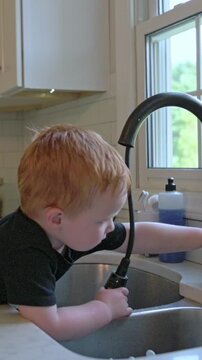 vertical screen redhaired boy tunes kitchen faucet flow, fairskinned child experimenting with water control at home sink, lighthaired boy adjusting tap to understand household plumbing functions