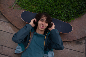 Smiling young man relaxing outdoors and listening music over a skateboard in a urban park