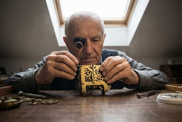 Elderly watchmaker repairing antique timepiece with loupe in dusty workshop