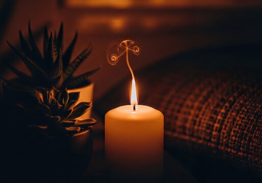 Warm glow of a burning candle with intricate smoke patterns by potted succulents in a dimly lit room - Powered by Adobe