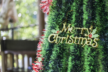 Joyful Merry Christmas gold sign decoration on festive green tree. Holiday greeting ornament for winter season celebration with shiny tinsel and beautiful blurred background