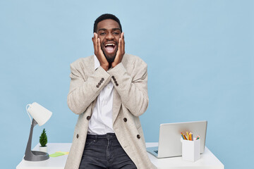 Joyful young man expressing excitement and surprise while sitting at a modern office desk with a laptop, stationery, and a desk lamp, featuring a plain blue background. People lifestyle concept