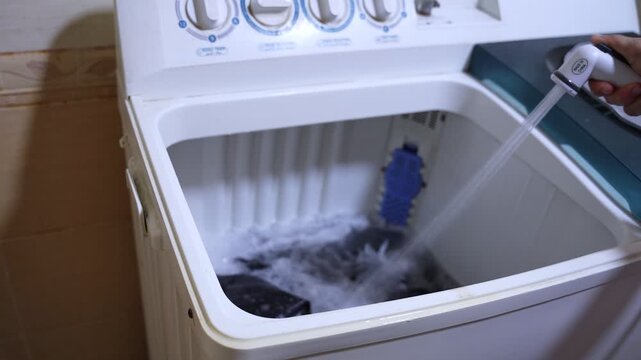 Close-up of a hand holding a spray nozzle to manually fill a top-loading washing machine with water. Authentic domestic laundry routine showing semi-automatic appliance use.