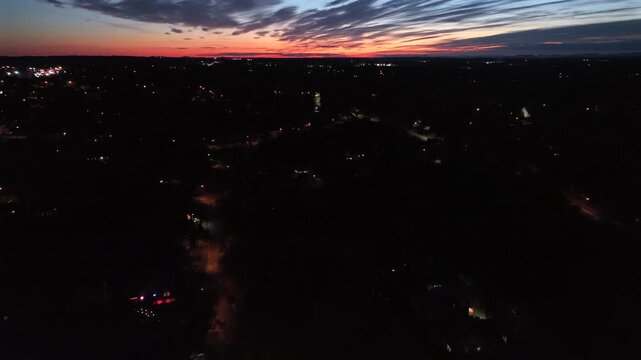 Aerial night view of an American town with scattered streetlights and houses glowing in dark. Dramatic sunset sky with vibrant orange and blue streaks on horizon. Wide shot. Virginia, USA.
