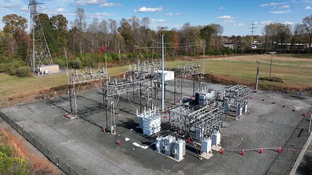 Aerial approaching shot of American electrical substation surrounded by open fields and autumn trees, showing metal structures, transformers and power lines under a clear blue sky.