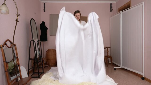 Barefoot woman energetically shaking out white, soft fleece throw blanket while standing at full length on bed in bedroom. Housewife is dressed in white long sleeved shirt and pink polka dot shorts