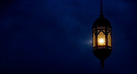 A glowing ornate lantern with intricate patterns hangs against a deep blue night sky. The warm light contrasts with the dark, evoking a serene, peaceful ambiance.