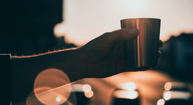 An intimate moment is captured as a person's hand delicately cradles a metallic cup against the soft glow of a sunset. The image evokes a sense of peace and contemplation.