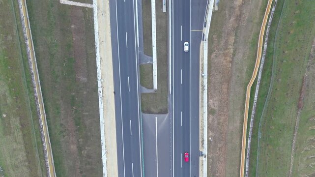 An aerial view of a multilane highway shows cars traveling on separate lanes. The road infrastructure, including barriers, markings, and surrounding grass areas, is clearly visible from above.