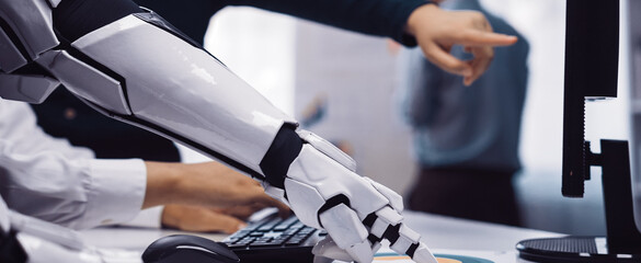 A futuristic robotic arm reaching towards a computer while human colleagues engage in discussion. This scene highlights teamwork, technology, and innovation in modern office environments.Noogenesis
