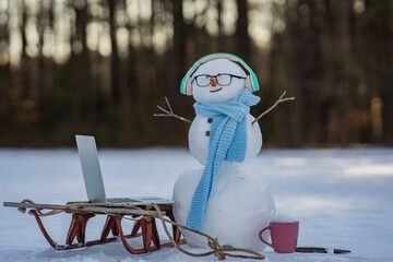 Snowman online freelancer scene in winter. Winter online shopping. Snowman doing remote work in winter landscape. Snowman with headphones and laptop in snow. Online shopping Christmas gift concept.