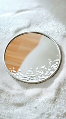 A round mirror with a broken section lies on a bed of white sand, reflecting a wooden surface. The image evokes themes of fragility and reflection.