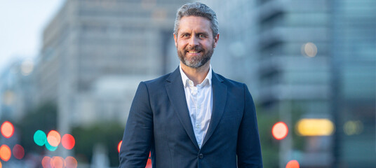 Happy mature businessman standing outside in an urban city street. Man in suit outdoor city night portrait. Mature business man walking on the city. Office worker posing on a city street.