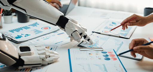 A futuristic robot hand interacts with human hands over business reports filled with charts and graphs, showcasing the fusion of technology and teamwork in modern workplaces.Noogenesis