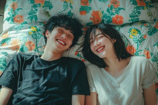 Lovely cute smiling young Asian lover couple lying on elbow on the bed and laughing while talking funny in the bedroom at home. Concept of romantic relationship.
