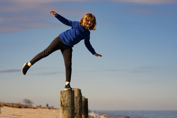 Child balances on wooden post. Kid plays near the beach. Kid keeps balance outdoors. Young boy playing on calm sea. Cute boy walking on a balance beam. Children on summer vacation. Happy childhood.