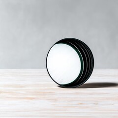 A circular object with black edges is resting on a wooden table, casting a shadow against a neutral gray background. The lighting is soft and even.