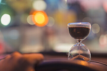 An hourglass placed on a car dashboard with night city bokeh lights symbolizes time pressure, focus, and decision-making. The warm, blurred ambiance enhances concepts of urgency, journey.