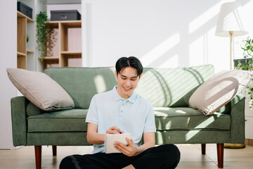 Young asian man relax on comfortable couch at home texting messaging on smartphone and tablet smiling man use cellphone  shopping online from home