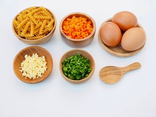 Pasta noodles are placed in a wooden bowl and chopped carrots, minced garlic, chopped spring onions and an  eggs on a white background.