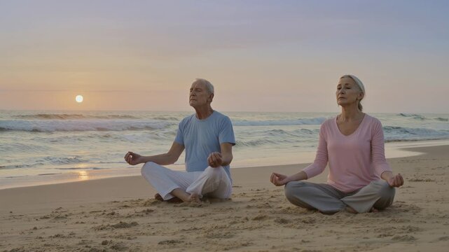 Un couple de seniors m&eacute;dite en position assise sur une plage au coucher du soleil, face &agrave; la mer