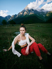 Obraz premium Mountains, landscape, outdoor, woman, meadow, grass, fashion, relaxed pose in a white crop top and red trousers resting on a lush meadow beneath blue sky and alpine peaks