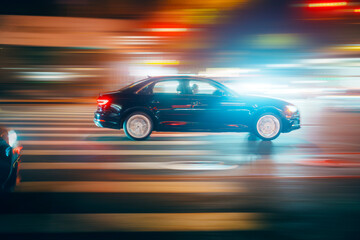 Black sedan car driving through dark street intersection in night city.. Long exposure. Bright colorful city lights blurred in background.