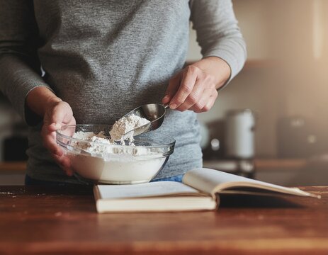 Woman baking in the kitchen, adding flour to a bowl while following a recipe from an open book
