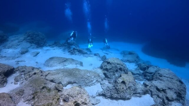 Group of scuba divers swimming in the ocean, sandy seabed with rocks, blue water. Ocean and scuba divers, underwater exploration video. Tourists in the sea.