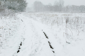 Narrow muddy path winding through a snowy field as snowflakes fall gently, creating a serene and peaceful winter landscape.