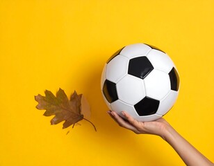 Autumn sports: Hand holding soccer ball with fallen leaf on yellow background