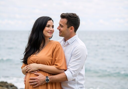 A loving pregnant couple smiling at each other on the beach. Man hugging his wife from behind and holding her belly. Expecting a baby concept - Powered by Adobe