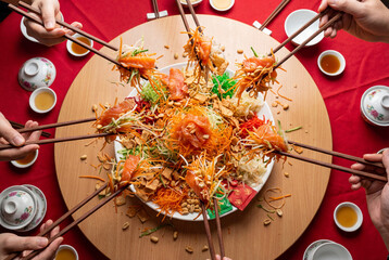 Obraz premium Multiple hands tossing salmon yusheng salad with chopsticks on a round table, festive shared meal for lunar new year celebration and family gathering tradition in Asian cuisine