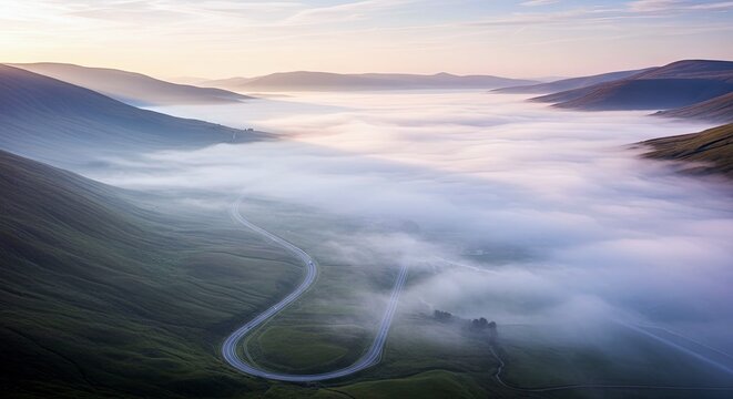 Aerial view of a road winding through green mountains, with a layer of fog or mist filling the valley at sunrise or sunset.