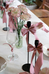 A beautifully set table features delicate pink carnations in a vase, elegant wine glasses adorned with pink ribbons, and black candle holders tied