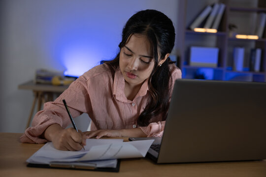 Asian young woman night work studying at desk writing notes with laptop open concentrating on task in dim home office with blue ambient light