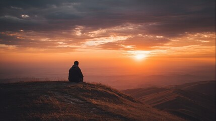 Fototapeta premium A person enjoys a quiet moment sitting on a hilltop observing the vibrant sunset. The sky displays warm colors as daylight gradually fades creating a peaceful atmosphere.