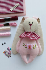 Handmade soft rabbit doll in pastel tones surrounded by pink craft supplies, delicate feminine vibe