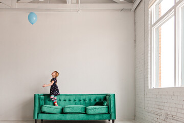 Girl standing on green sofa while watching blue balloon float indoors