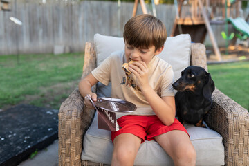 Boy sitting on chair and eating a s'mores with a dachshund next to him