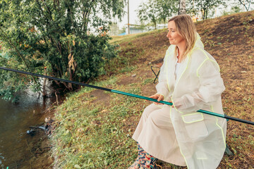 woman sits by river, fishing, rain. white raincoat. trees and house