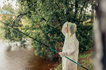 woman stands by river, fishing, rain. white raincoat. trees and house