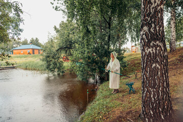 woman stands by river, fishing, rain. white raincoat. trees and house