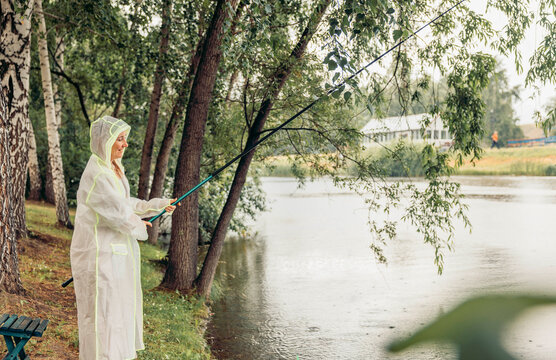 woman stands by river, fishing, rain. white raincoat. trees and house