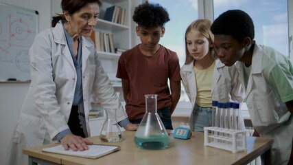 Woman teacher with multiracial kids in lab coat observing blue chemical reaction in flask during science experiment. Early education concept.